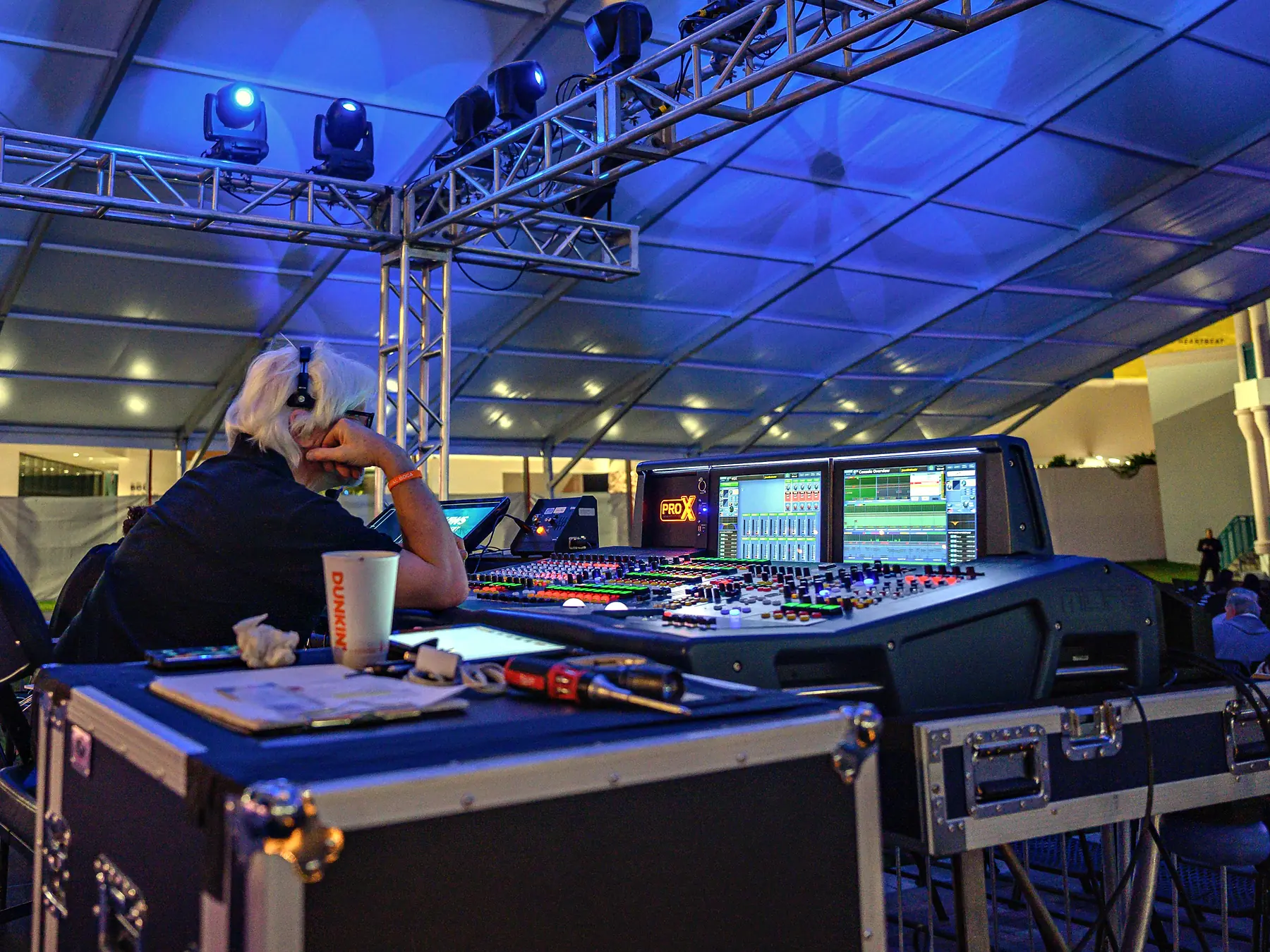 An audio engineer in headphones at a mixing console under a tent roof washed blue, a truss of moving lights overhead and the open side of the tent beyond.