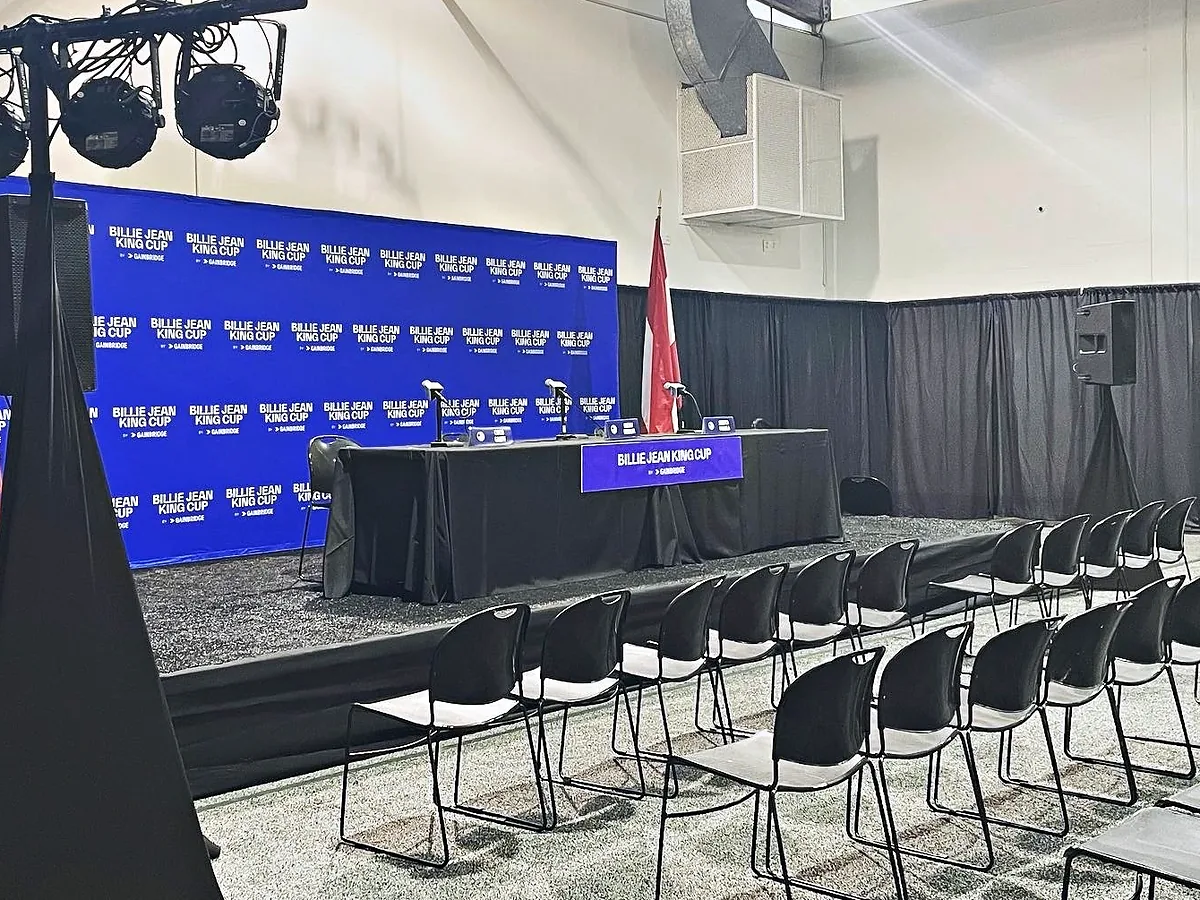 A skirted head table with microphones on a low riser in front of a branded step-and-repeat backdrop, a lighting tree at the left, a loudspeaker on a stand at the right, a flag beside the table and rows of empty chairs facing it.