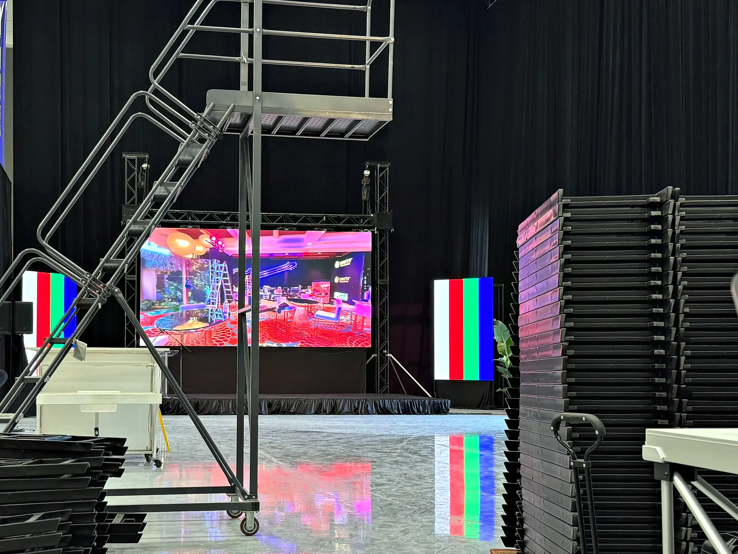 An LED wall standing on ground support behind a skirted deck in a black-draped room, a rolling stair tower beside it, two narrow panels driven to color test fields, and stacked folding furniture on carts to either side.