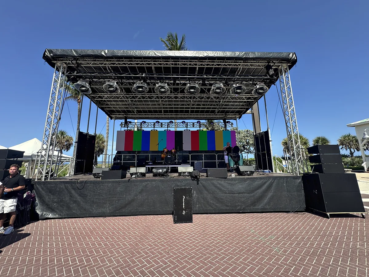 A covered outdoor stage on a brick plaza: a trussed roof with a row of lighting fixtures under it, loudspeakers stacked either side of the deck, and an LED backdrop driven to color test fields against palms and a clear sky.