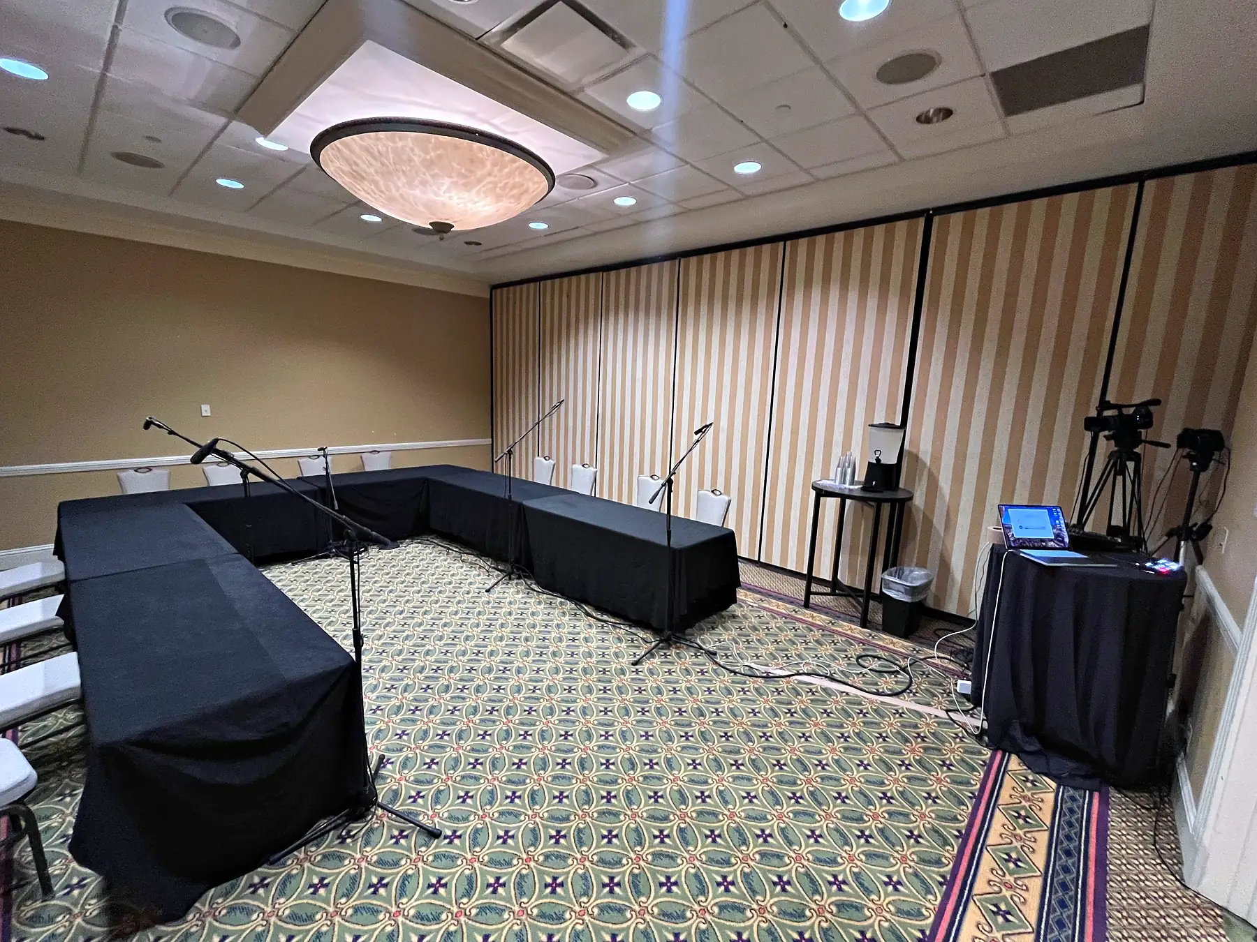 A meeting room with black-skirted tables and white chairs, microphones on boom stands along them, and a camera on a tripod and a laptop on a draped table at the side of the room.