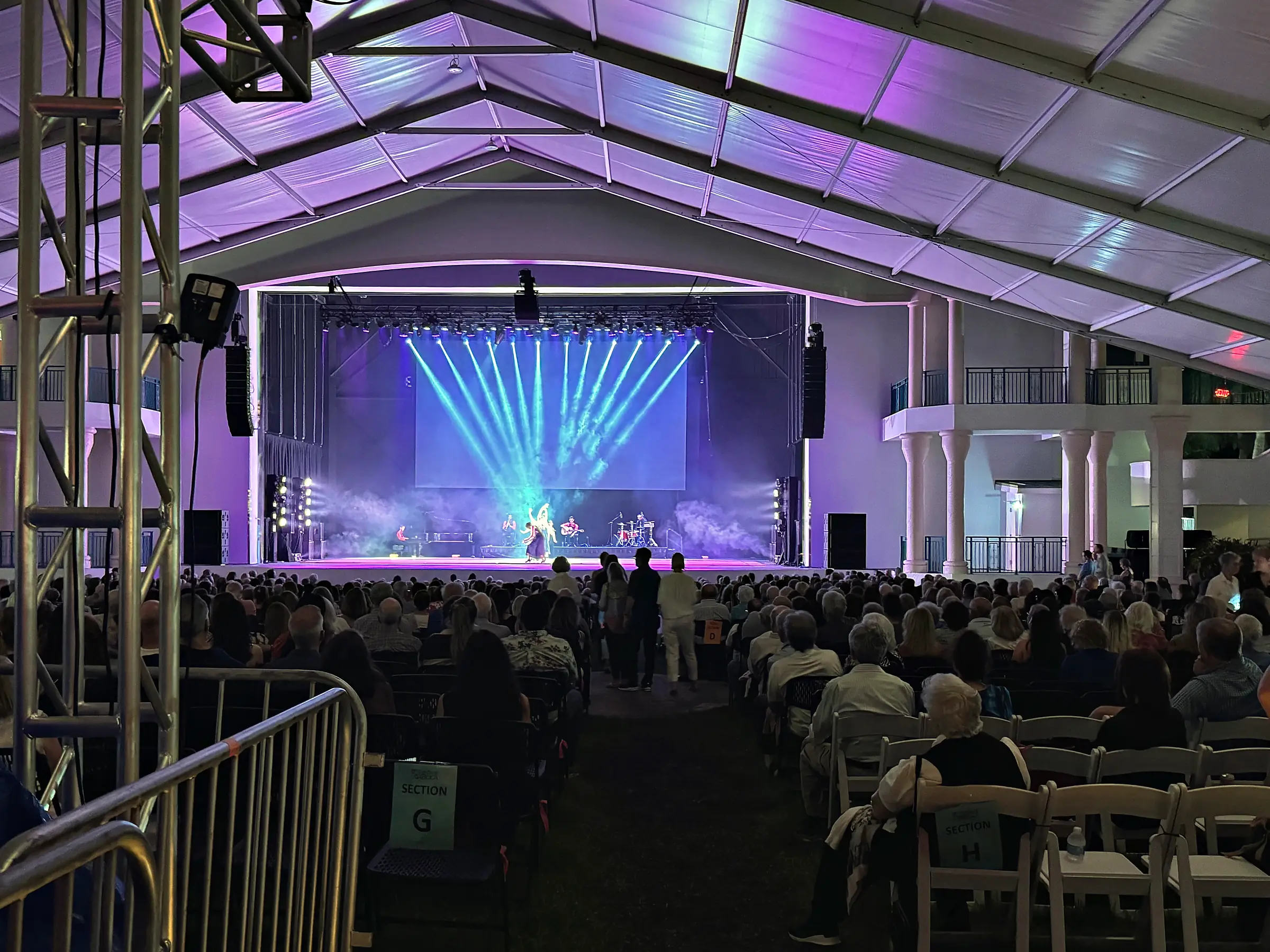Rows of seated guests under a tent roof washed purple, facing a lit stage with beams of light fanned out above it, and a truss tower in the foreground at the left.