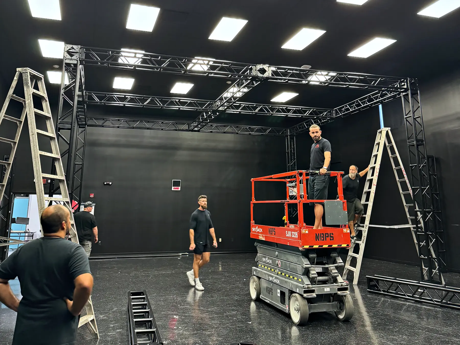 A box of ground-supported truss standing in a black-walled room, a scissor lift under it with a crew member on the platform, stepladders to either side and more of the crew on the floor.